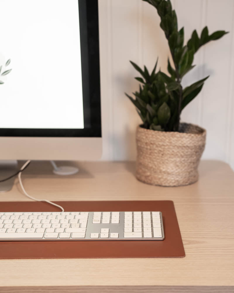 DESK skrivunderlägg i cognac läder med tangentbord framför iMac och grön växt i korg på ljust skrivbord