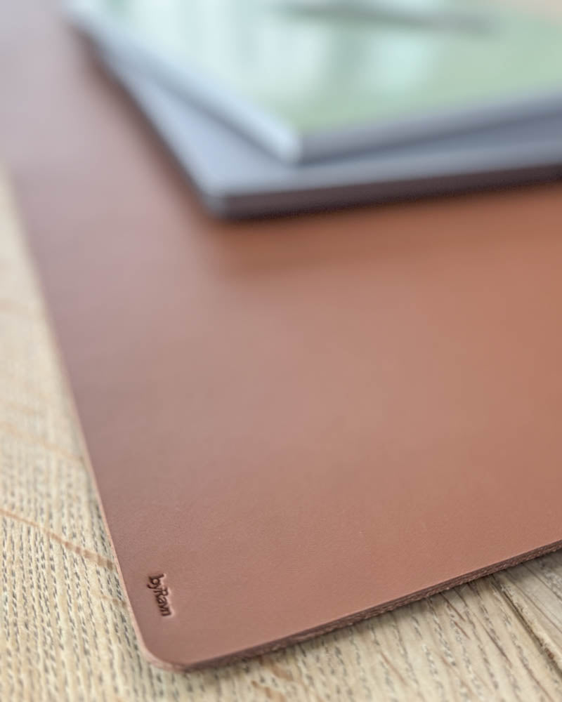 DESK skrivunderlägg i cognac läder, närbild av hörnet med byRavn-prägling under laptop på träbord