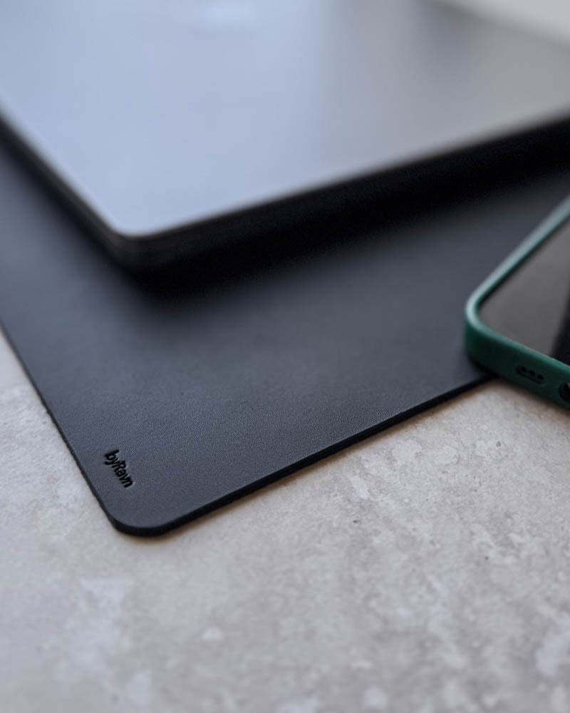 DESK skrivunderlägg i svart läder, närbild av hörnet med byRavn-prägling under laptop och telefon