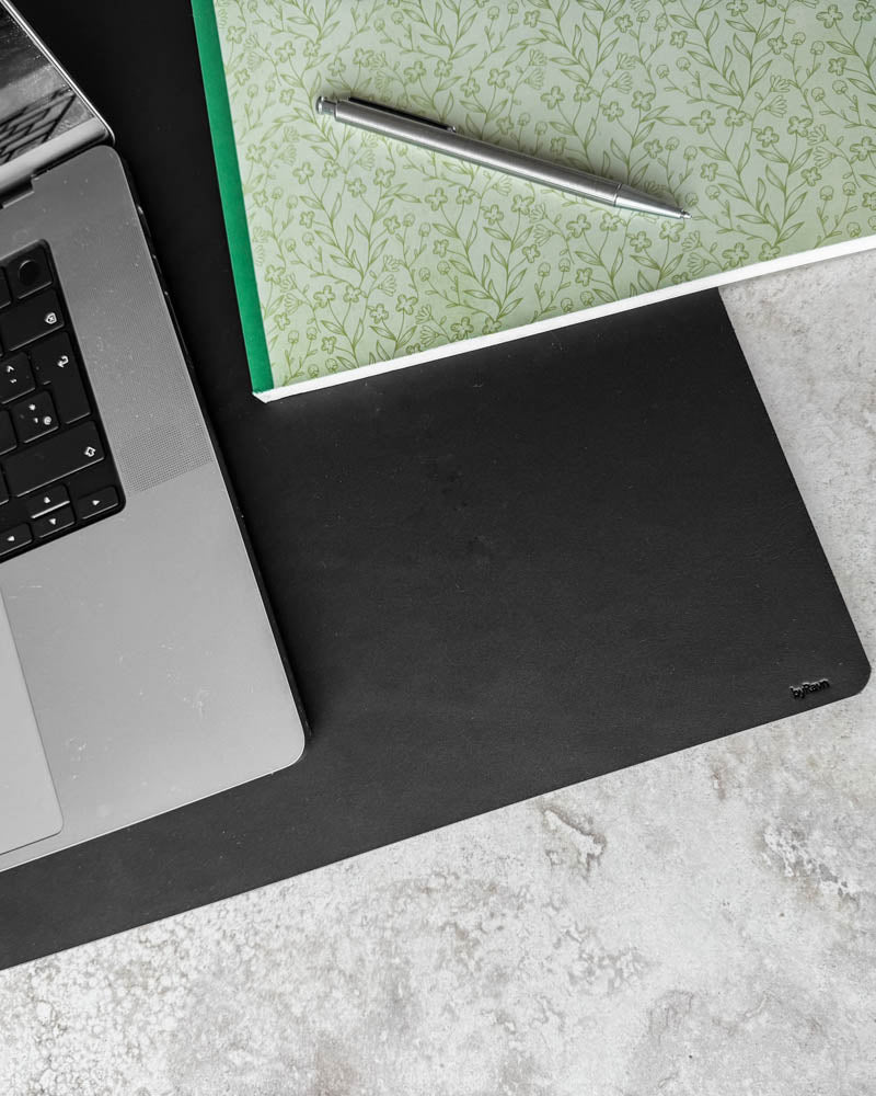 DESK skrivunderlägg i svart läder sett ovanifrån med laptop, blommig anteckningsbok och kulspetspenna på ljus bordsskiva
