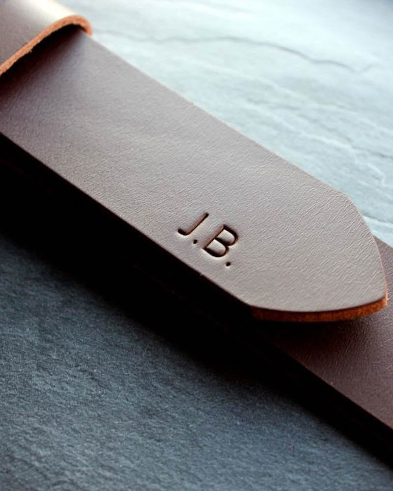 EDGE bälte i mörkbrunt läder i närbild med initialerna "J.B." graverade på spetsen, på skifferplatta