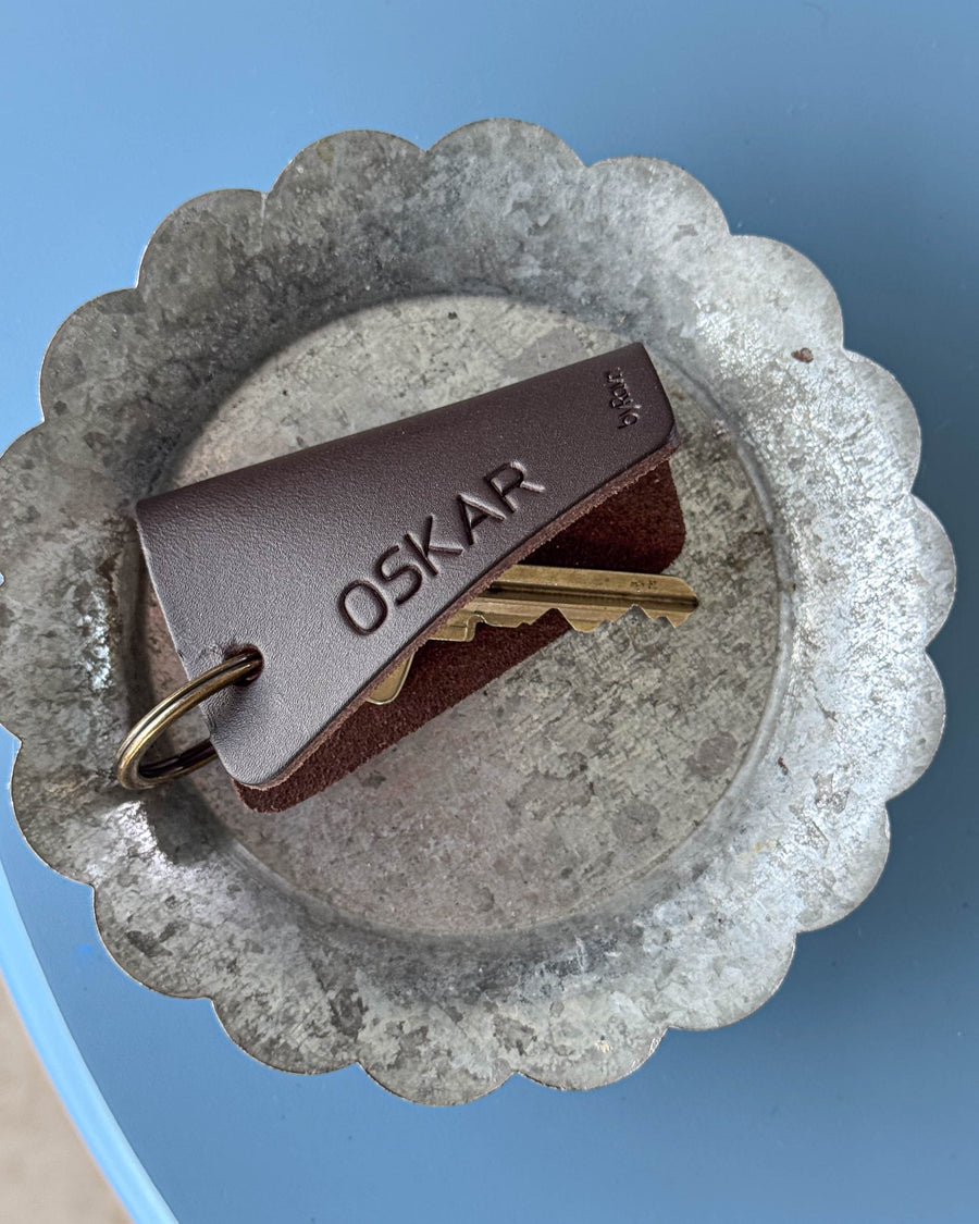 FOLD nyckelfodral i mörkbrunt läder med namnet "Oskar" graverat, med nycklar i zinkfat på blått bord