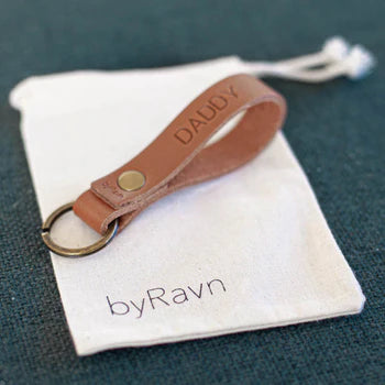 Presentpåse i vitt tyg med byRavn-logotyp och nyckelring i cognac läder med gravyren "Daddy" på grönt underlag