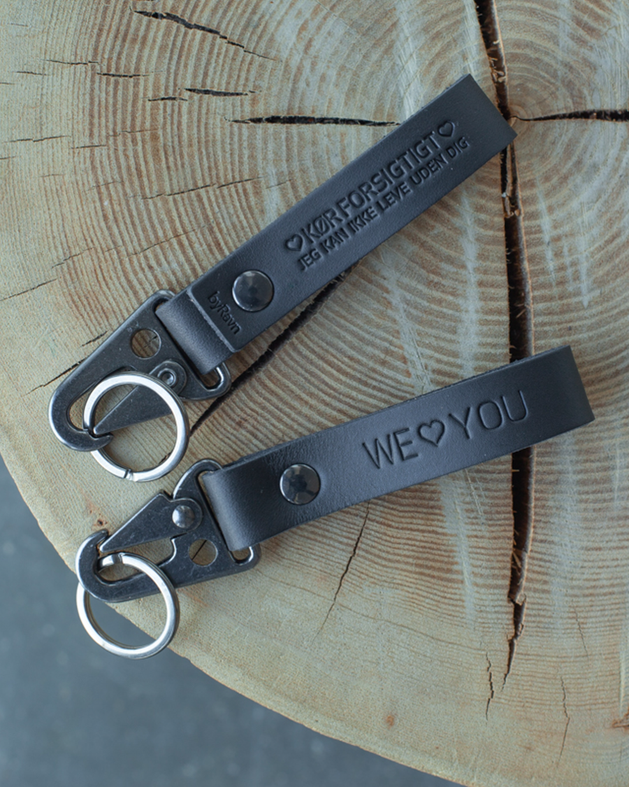 Två HOOK nyckelringar i svart läder på trästubbe med gravyrerna "We ♡ you" och "Kør forsigtigt – jeg kan ikke leve uden dig"