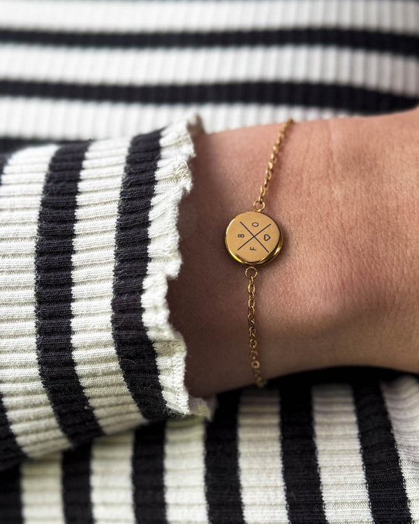 IBI X armband i guldpläterat stål med kryss, bokstäver och hjärta graverade, buret runt handleden till randig blus