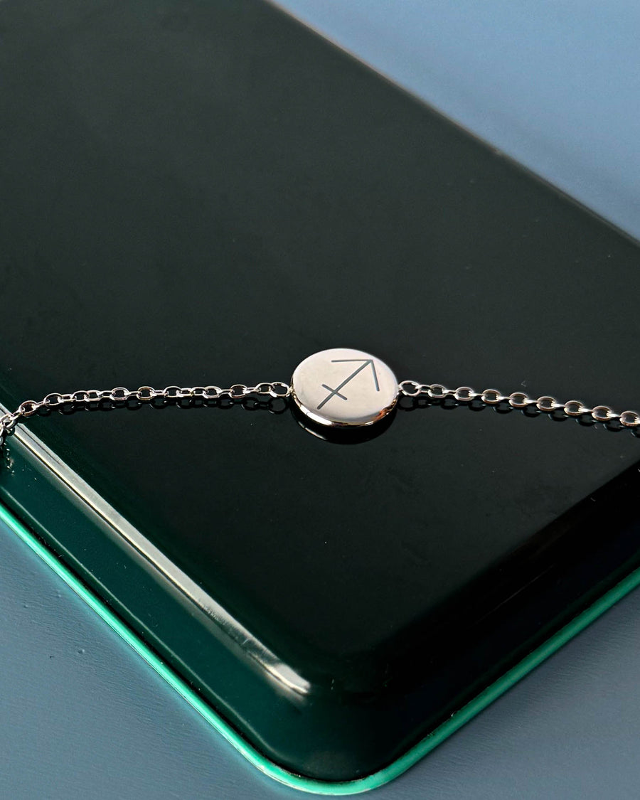 IBI STAR armband i silverfärgat rostfritt stål med stjärntecknet Skytten graverat på rund bricka, liggande på mörkgrön ask