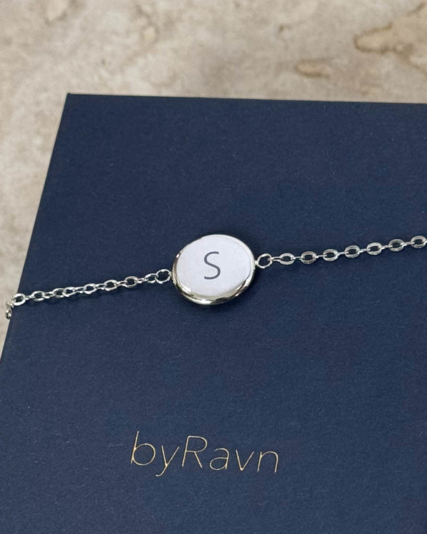 IBI LETTER armband i silverfärgat rostfritt stål med bokstaven "S" graverad, liggande på mörkblå byRavn smyckesask