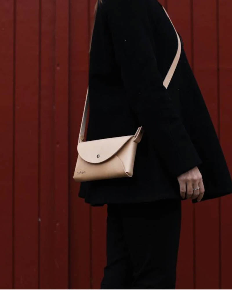 INA axelväska i naturfärgat läder buren crossbody av kvinna i svarta kläder framför röd träpanel