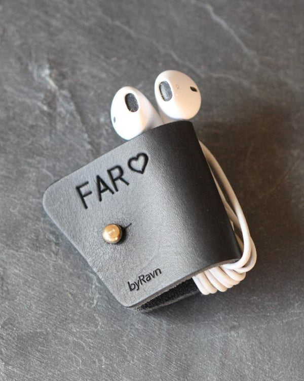 KAX hörlurshållare i svart läder med personlig gravyr "Far" ♡ och mässingsknapp, med earpods på mörk skiffer