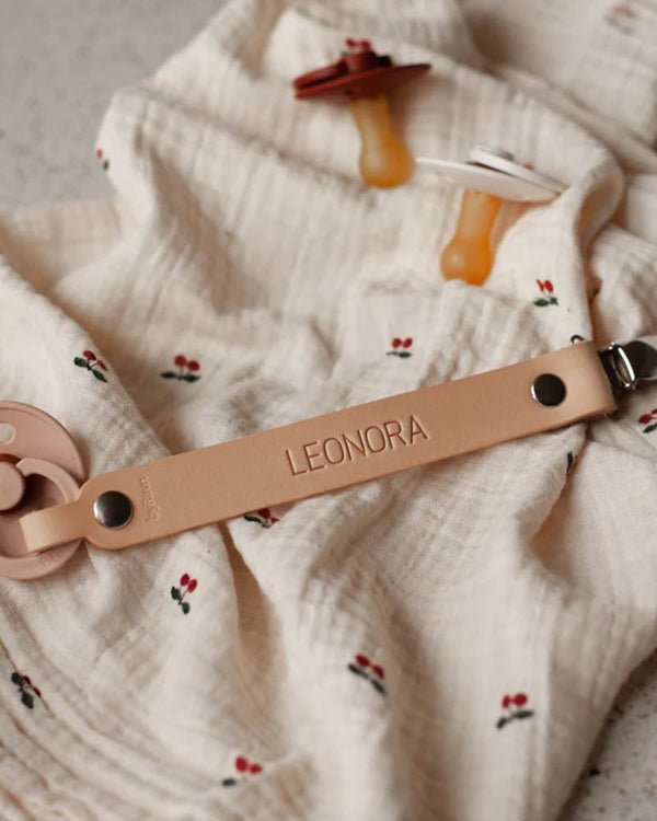 KODA nappband i naturfärgat läder med namnet "Leonora" graverat, lagt på tygblöja med körsbär och två nappar
