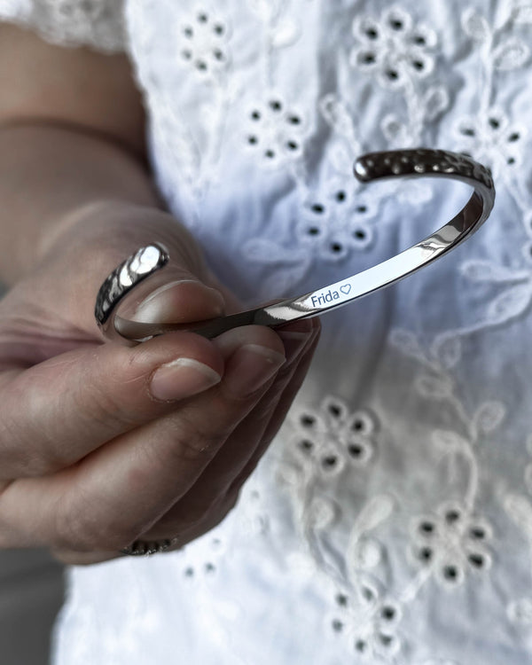 LUNA armband i silverfärgat rostfritt stål som hålls i handen, med namnet "Frida" ♡ graverat på insidan