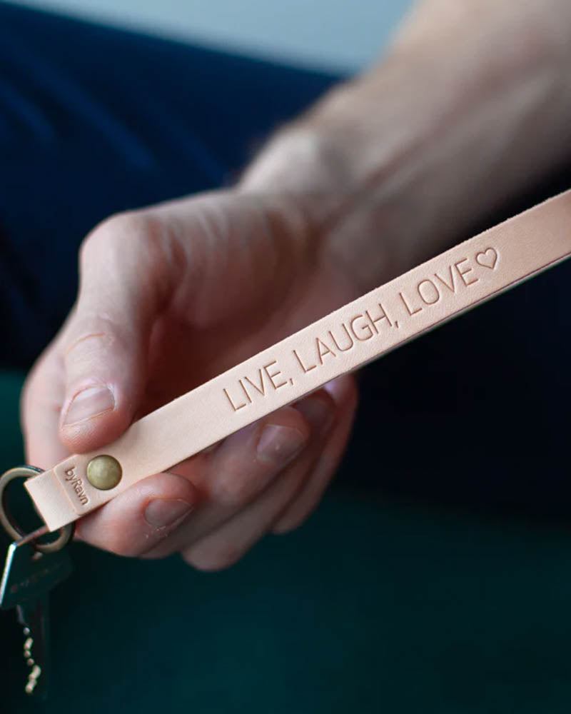 LUX nyckelring i naturfärgat läder med mässingsnit och personlig gravyr "Live, laugh, love" ♡, hålls i handen med nyckel