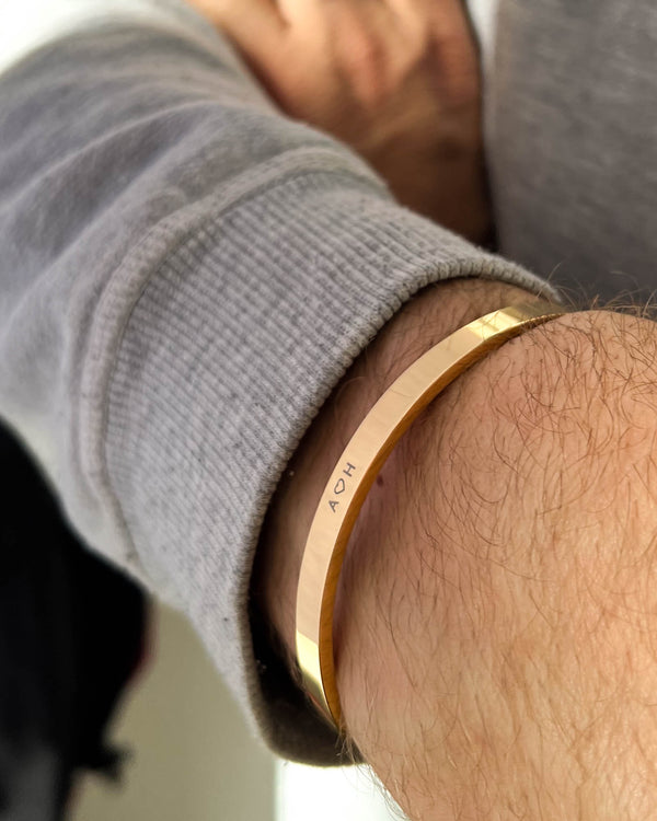 MARS armband i guldpläterat stål med initialerna "A ♡ H" graverade runt manshandled, grå sweatshirt
