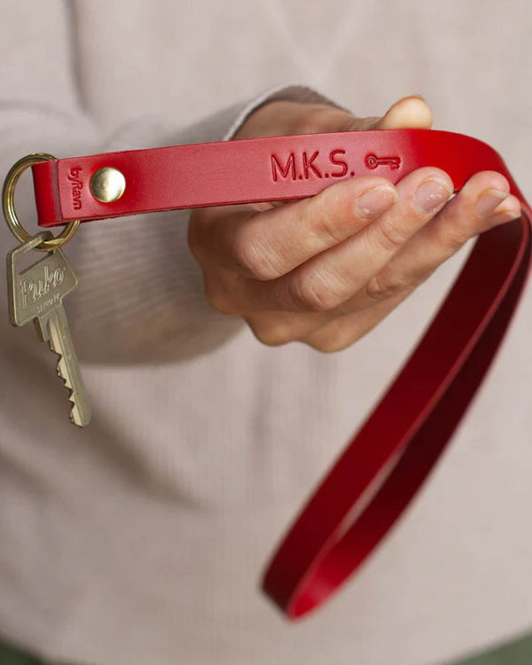 MAX nyckelring i rött läder som lång stropp med initialerna "M.K.S." och nyckelsymbol graverade, hålls i handen med nyckel