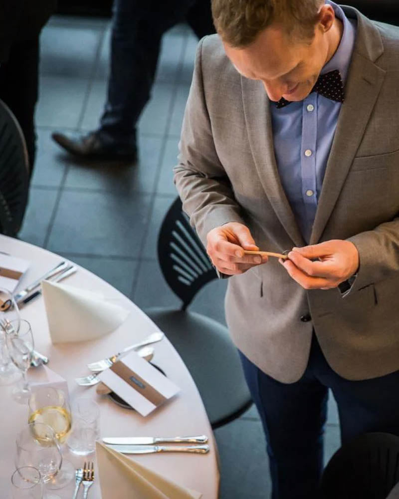 MERRY placeringskort i läder hålls i handen av festklädd gäst i kostym och fluga vid dukat runt bord