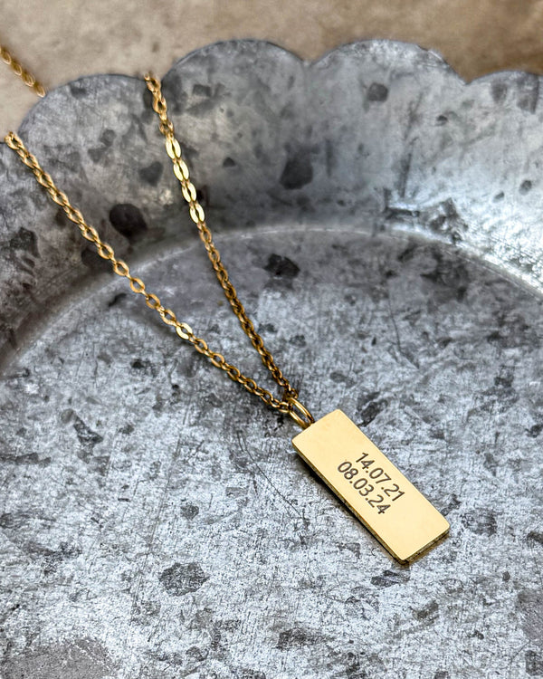 MIO halsband i guldpläterat stål med datumen "14.07.21 08.03.24" graverade på rektangulärt hänge, på zinkfat med vågig kant