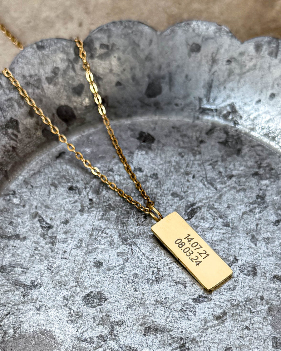 MIO halsband i guldpläterat stål med datumen "14.07.21 08.03.24" graverade på rektangulärt hänge, på zinkfat med vågig kant