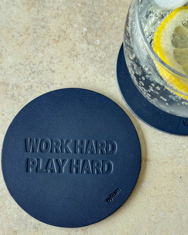 MOKKA glasunderlägg i mörkblått läder präglat "Work hard play hard" på ljust bord vid glas med citronskiva