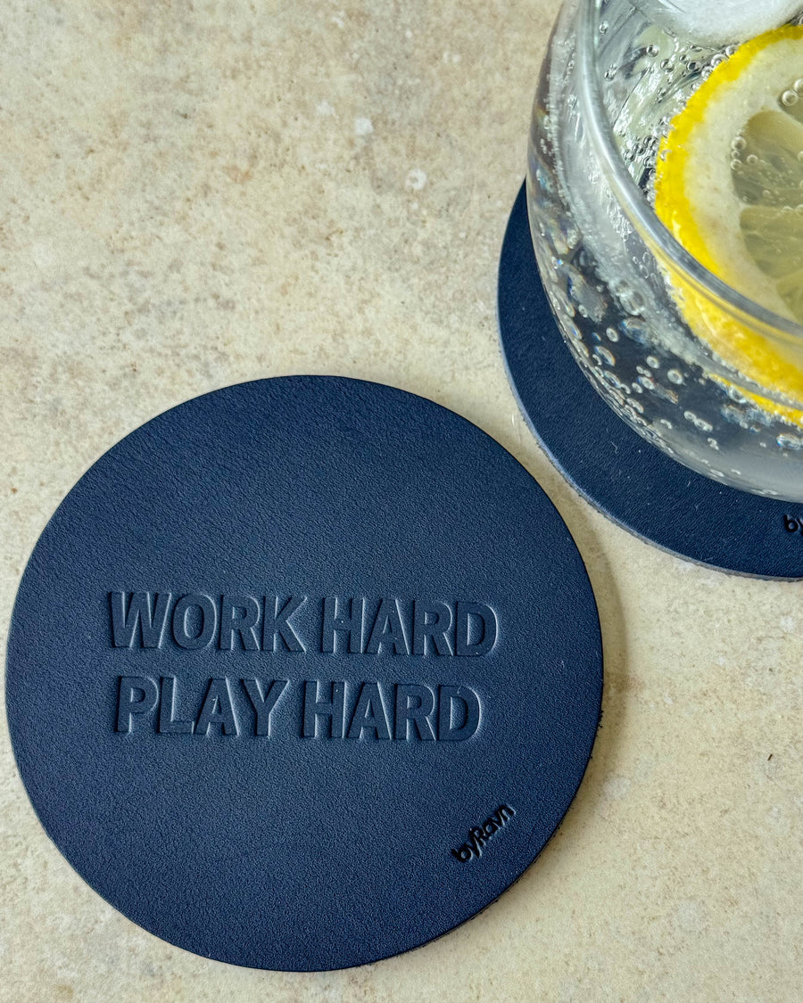 MOKKA glasunderlägg i mörkblått läder präglat "Work hard play hard" på ljust bord vid glas med citronskiva