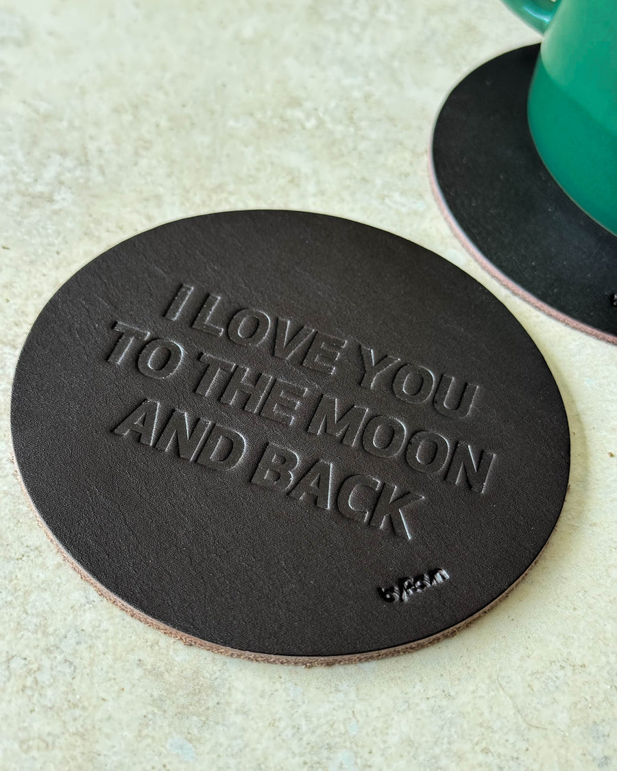 MOKKA glasunderlägg i mörkbrunt läder präglat "I love you to the moon and back" på ljust bord vid grön kopp