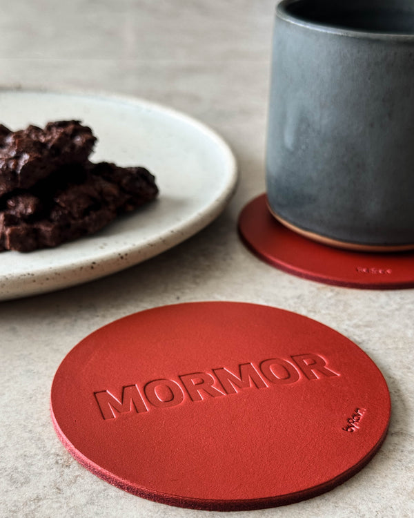 MOKKA glasunderlägg i rött läder präglat "Mormor" på ljust bord med grå kaffekopp och tallrik med chokladkaka