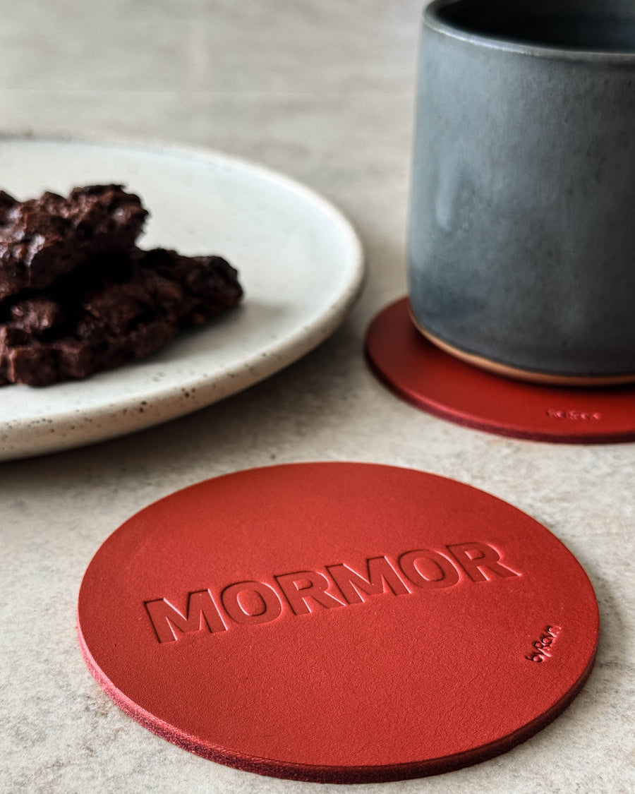 MOKKA glasunderlägg i rött läder präglat "Mormor" på ljust bord med grå kaffekopp och tallrik med chokladkaka