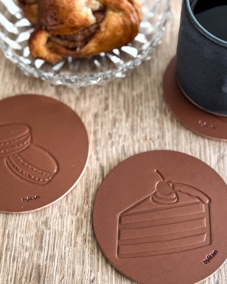 MOKKA kaffe & kaka-glasunderlägg i cognacfärgat läder präglade med tårta och macarons på träbord vid kanelbulle och svart kopp