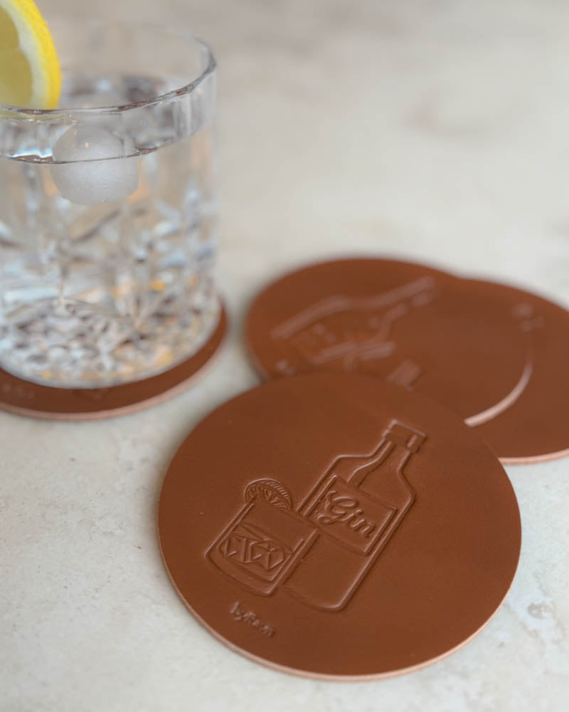 MOKKA sprit-glasunderlägg i cognacfärgat läder präglade med ginflaska och drink, på ljus stenyta vid glas med citronskiva