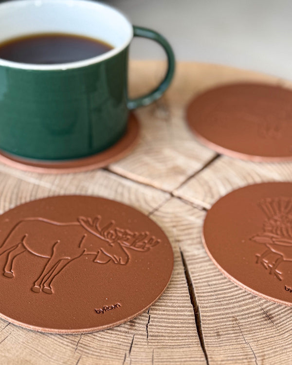 MOKKA jakt-glasunderlägg i cognacfärgat läder präglade med älg och tjäder på träskiva vid grön kaffekopp på underlägg