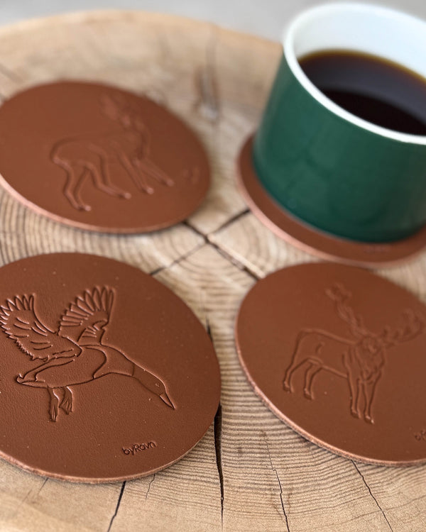 MOKKA jakt-glasunderlägg i cognacfärgat läder präglade med flygande gräsand, råbock och kronhjort på träskiva med grön kaffekopp