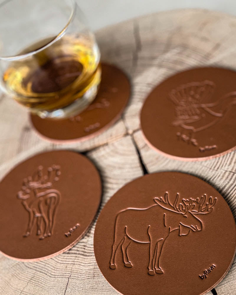 MOKKA jakt-glasunderlägg i cognacfärgat läder präglade med älg, dovhjort och tjäder på träskiva vid glas med whisky