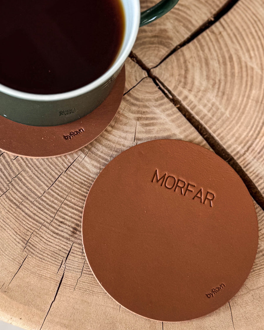MOKKA runt glasunderlägg i cognacfärgat läder med gravyren "Morfar" på träskiva bredvid grön kaffekopp på underlägg