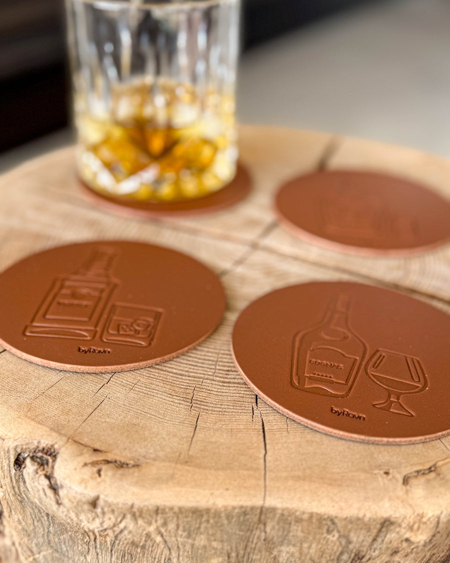 MOKKA sprit-glasunderlägg i cognacfärgat läder präglade med whisky och cognac på träskiva vid kristallglas med whisky och is