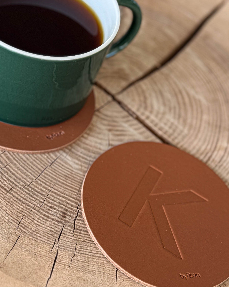 MOKKA glasunderlägg i cognacfärgat läder präglat med bokstaven "K" på träskiva vid grön kaffekopp på underlägg