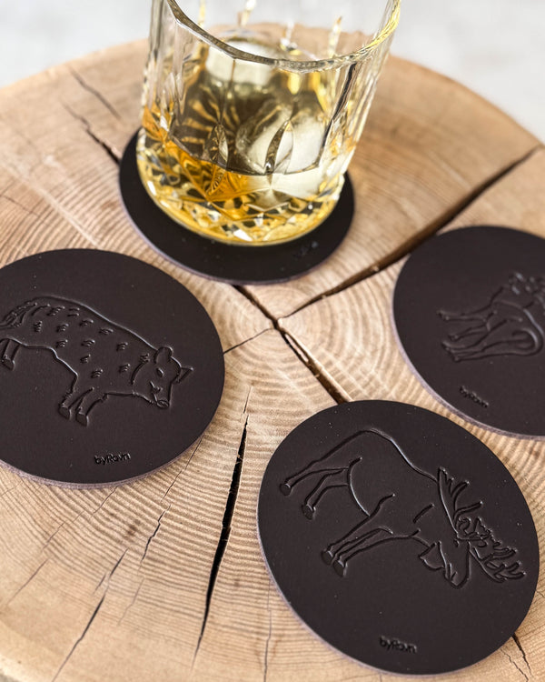 MOKKA jakt-glasunderlägg i mörkbrunt läder präglade med vildsvin, älg och dovhjort på träskiva vid whiskyglas