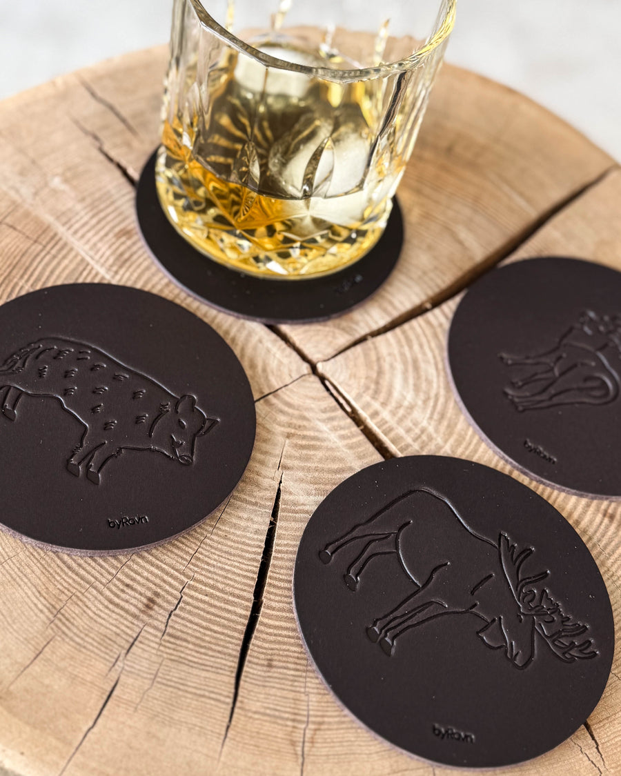 MOKKA jakt-glasunderlägg i mörkbrunt läder präglade med vildsvin, älg och dovhjort på träskiva vid whiskyglas