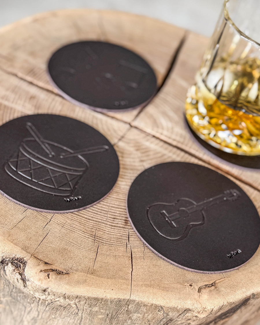 MOKKA glasunderlägg i mörkbrunt läder präglade med trumma och gitarr på träskiva vid kristallglas med whisky