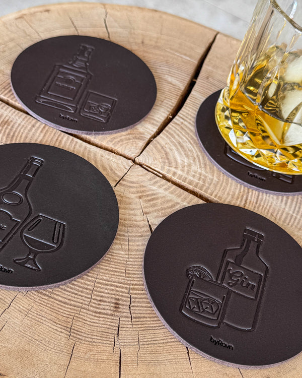 MOKKA sprit-glasunderlägg i mörkbrunt läder präglade med gin, cognac och whisky på träskiva vid glas med whisky och is
