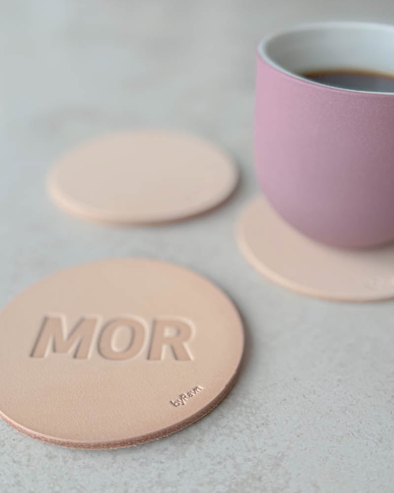 MOKKA glasunderlägg i naturfärgat läder präglat "Mor" i stora bokstäver, rosa kaffekopp på underlägg bakom