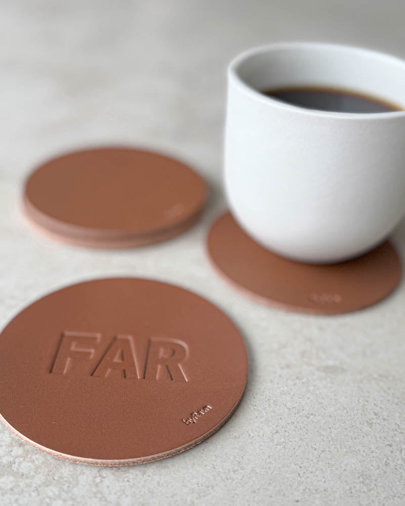 MOKKA glasunderlägg i cognacfärgat läder präglat "Far" i stora bokstäver, fler underlägg och vit kaffekopp bakom