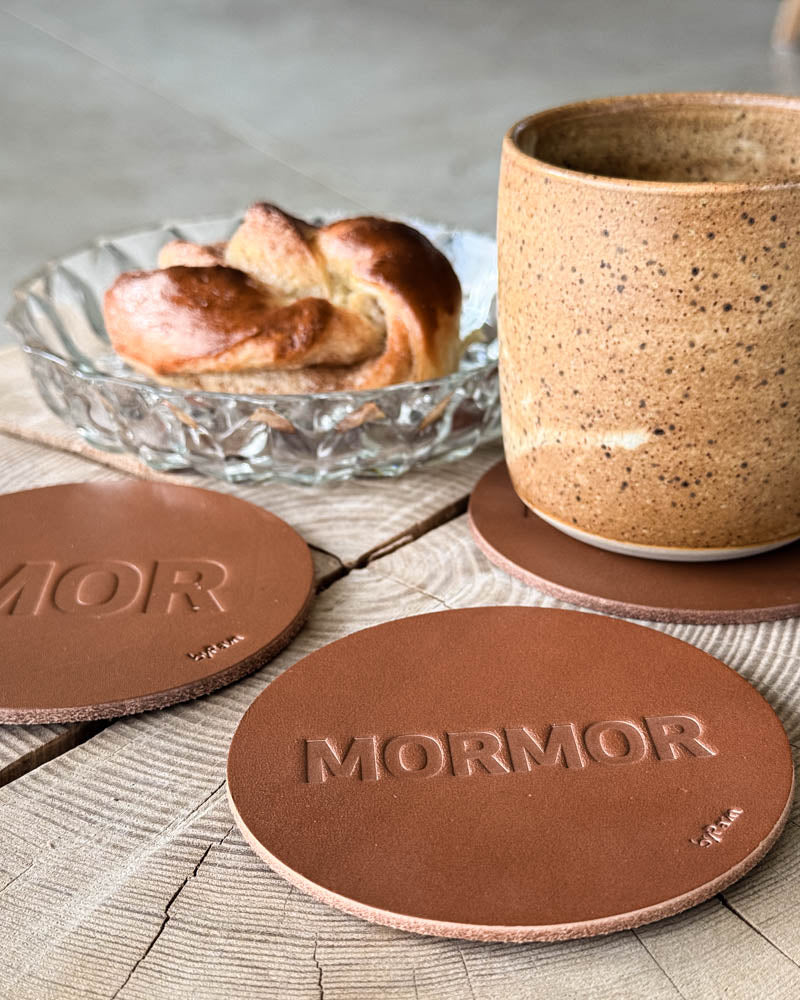 MOKKA glasunderlägg i cognacfärgat läder präglade "Mor" och "Mormor" på träbord med kaffekopp och kanelbulle i glasskål