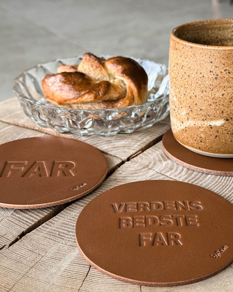 MOKKA glasunderlägg i cognacfärgat läder präglade "Far" och "Verdens bedste far" vid kaffekopp och kanelbulle på träbord