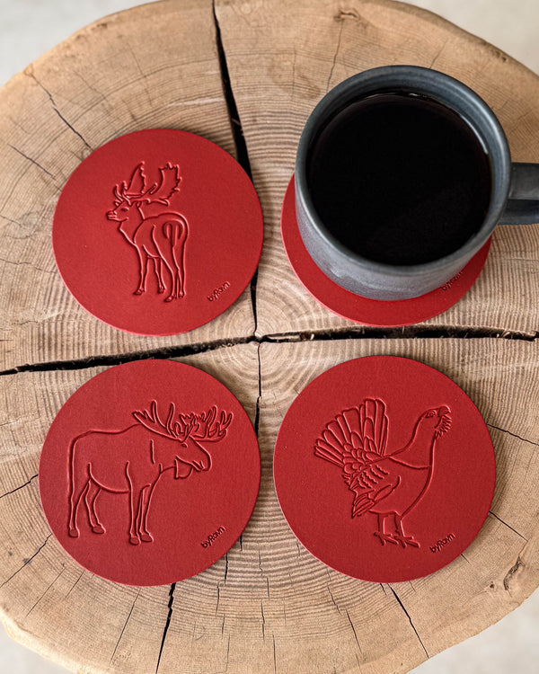 MOKKA jakt-glasunderlägg i rött läder präglade med älg, tjäder och dovhjort på träskiva vid grå kaffekopp på underlägg