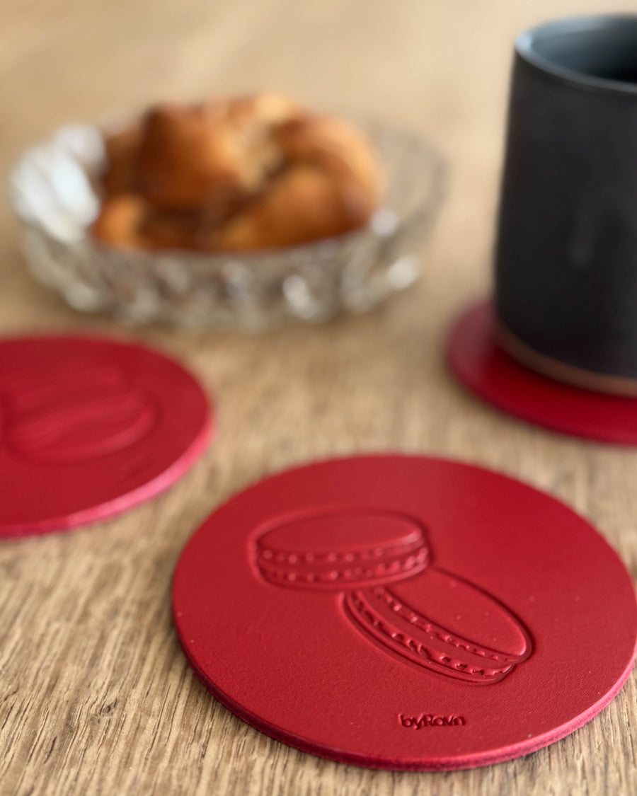 MOKKA kaffe & kaka-glasunderlägg i rött läder präglat med macarons på träbord vid svart kopp och kanelbulle i glasskål