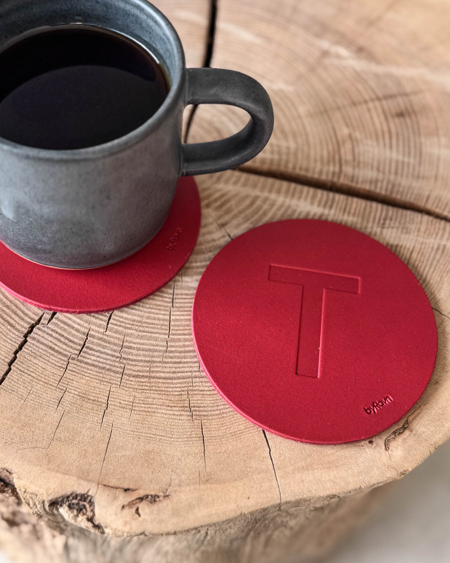 MOKKA glasunderlägg i rött läder präglat med bokstaven "T" på träskiva vid grå kaffekopp på underlägg