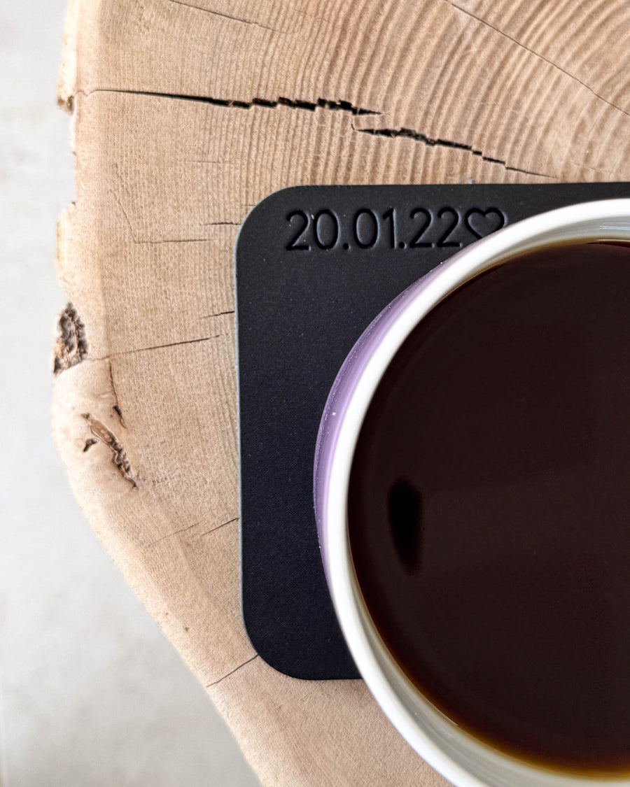 MOKKA fyrkantigt glasunderlägg i svart läder med personlig gravyr "20.01.22" ♡ under kaffekopp på träskiva