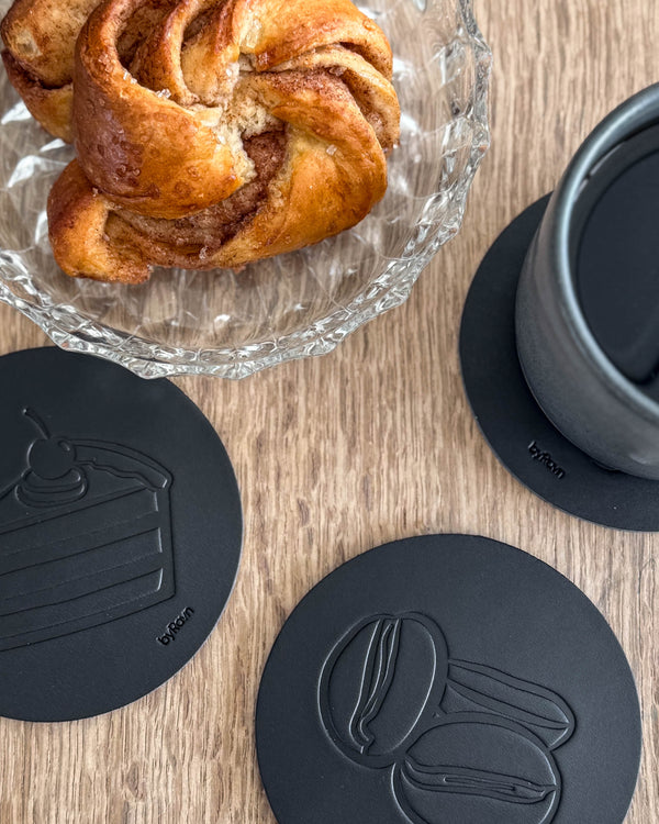 MOKKA kaffe & kaka-glasunderlägg i svart läder präglade med tårta och kaffebönor på träbord vid kanelbulle och grå kopp