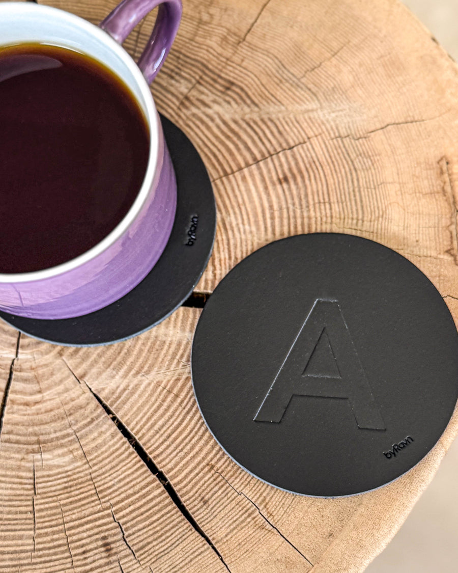 MOKKA glasunderlägg i svart läder präglat med bokstaven "A" på träskiva vid lila kaffekopp på underlägg