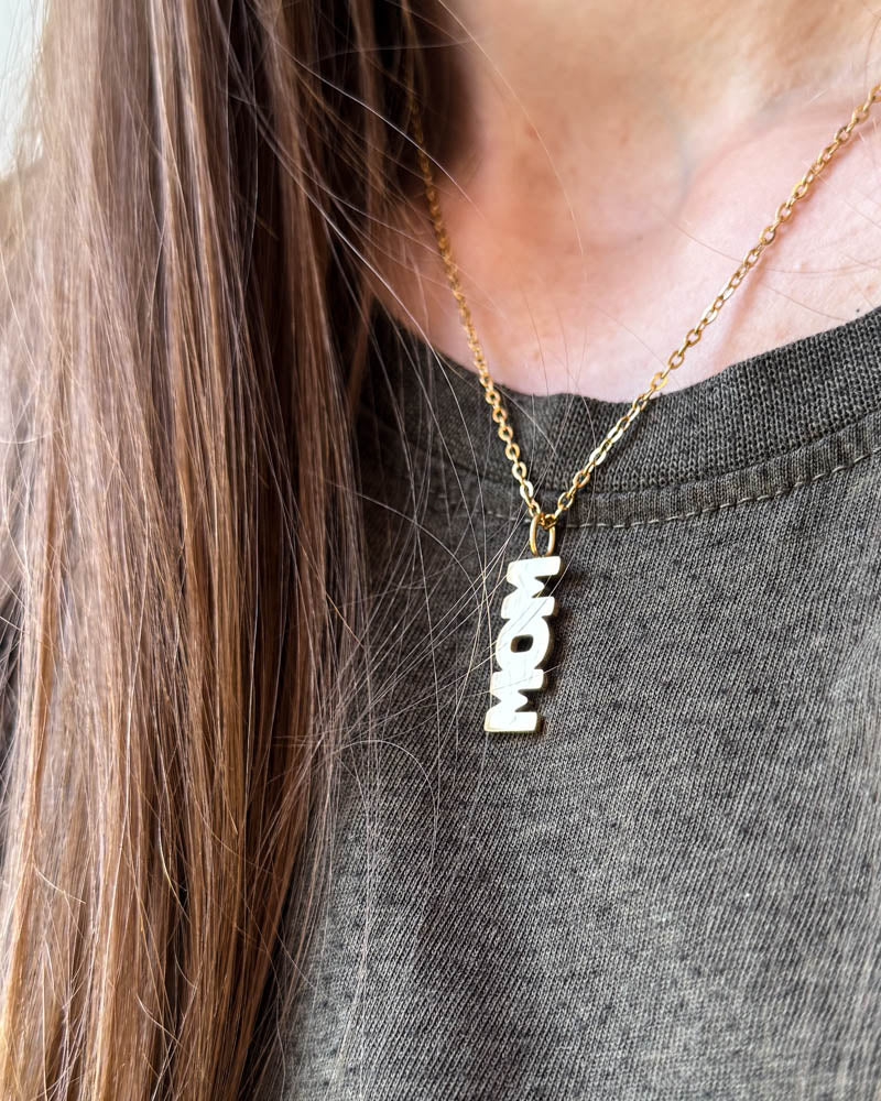 MOM halsband med bokstavshänget "MOM", här visat i guldpläterat stål, buret runt halsen över grå t-shirt