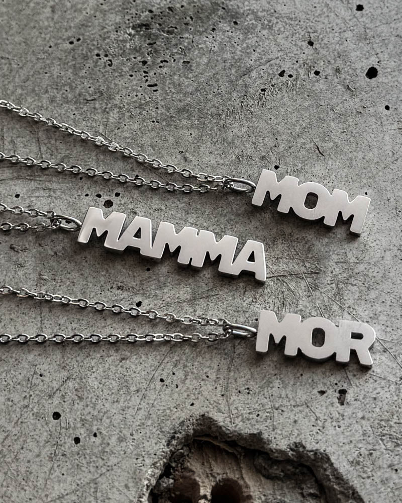 MOM halsband i silverfärgat rostfritt stål med bokstavshängen "MOM", "MAMMA" och "MOR", liggande på rå betong